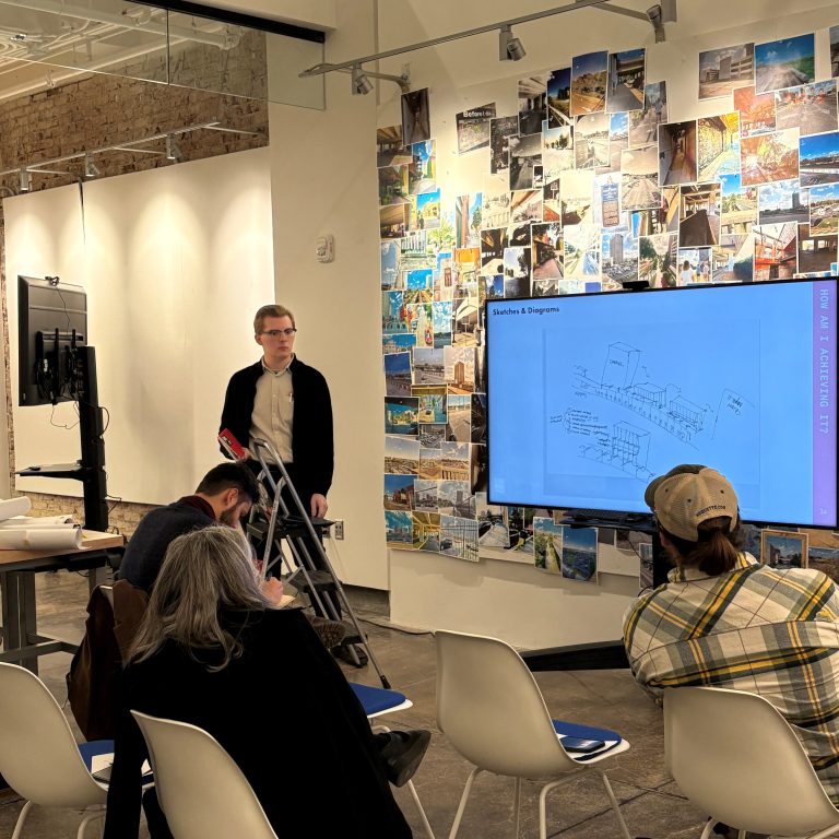 A person stands with crutches giving a presentation to seated attendees, with a screen displaying diagrams and notes in a modern room decorated with numerous photos on the wall.