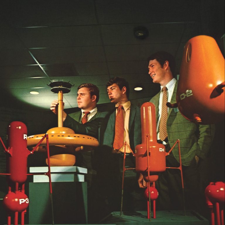 Three men in suits examine red and orange industrial models displayed on pedestals at an indoor ARMCO National Supply Division exhibition.