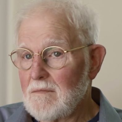 Elderly man with white hair, beard, and glasses wearing a blue collared shirt and striped t-shirt, looking directly at the camera.