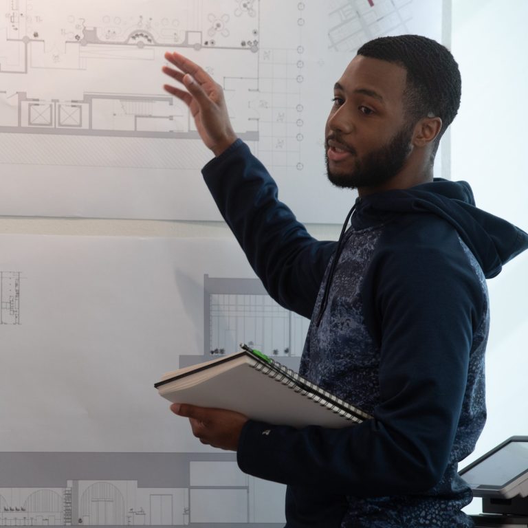 A man stands in front of architectural drawings on the wall, holding a notebook and gesturing while speaking to another person.
