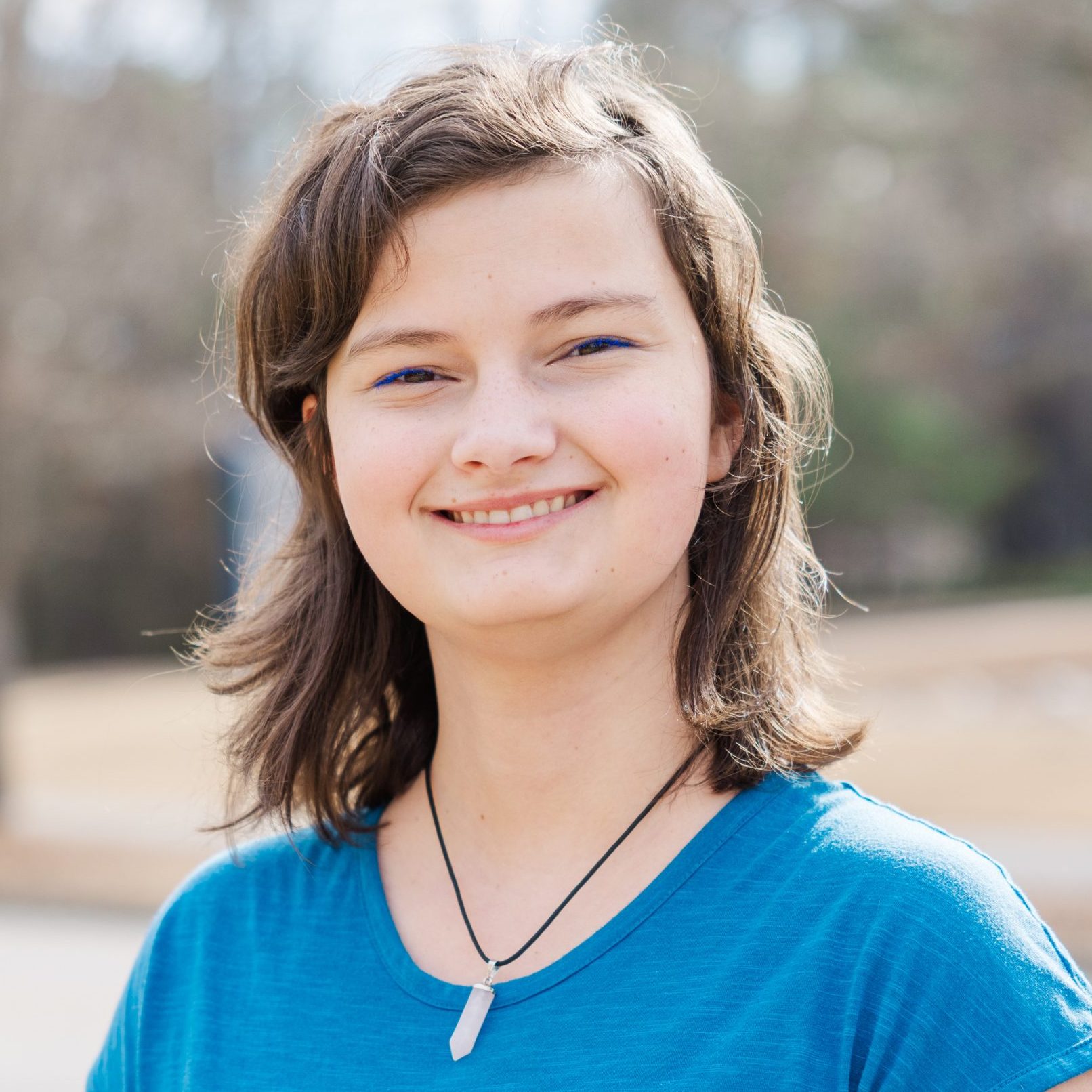 A person with wavy brown hair wearing a blue shirt and a pendant necklace smiles outdoors on a sunny day.