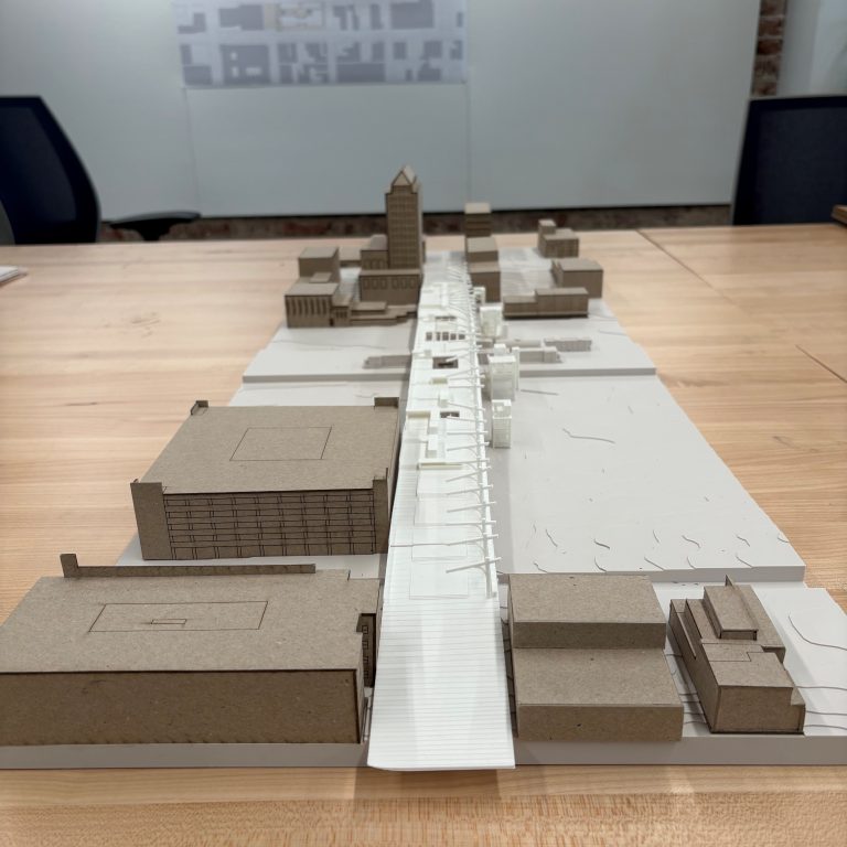 A tabletop architectural model of an urban area, showing buildings made of cardboard and white material, with a central street running through the scene.
