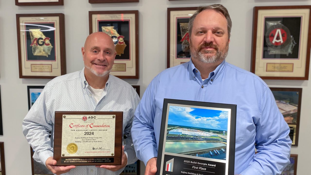 Two men pose indoors in front of a wall of plaques, each holding an award; one is a certificate, the other a framed photo with a “First Place” construction award.