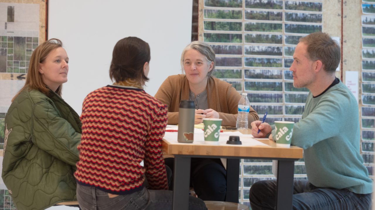 Four adults sit around a table engaged in conversation, with papers, mugs, and a water bottle on the table. A wall with photos is visible in the background.