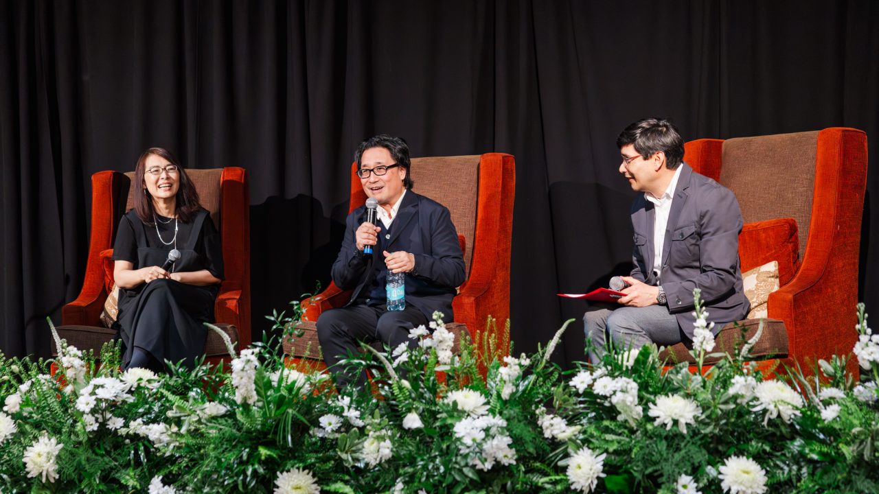 Three people sit on stage in large chairs, speaking and laughing, with floral arrangements in front and a black curtain backdrop.
