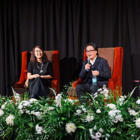 Three people sit on stage in large chairs, speaking and laughing, with floral arrangements in front and a black curtain backdrop.