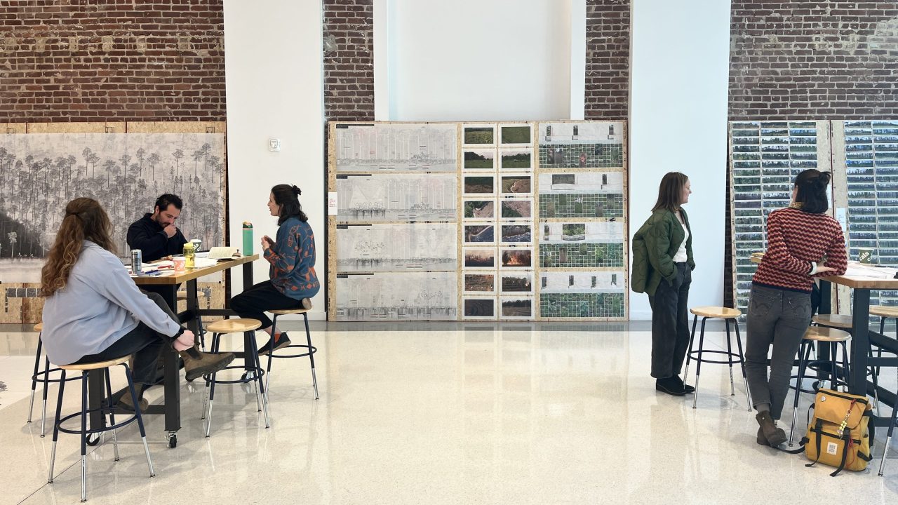 Six people work and converse in a bright room with brick walls, large artworks, and high ceilings; some sit at tables while others stand and talk.