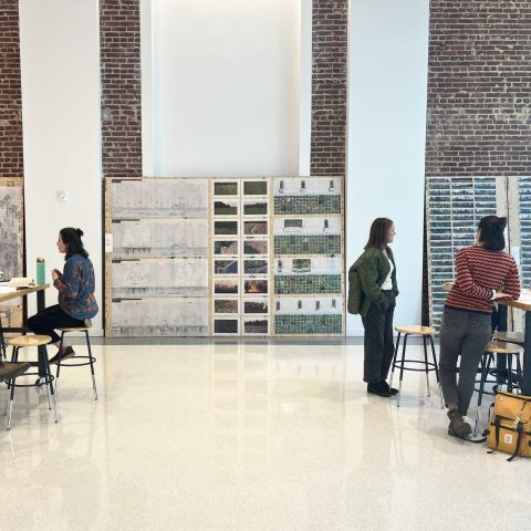 Six people work and converse in a bright room with brick walls, large artworks, and high ceilings; some sit at tables while others stand and talk.