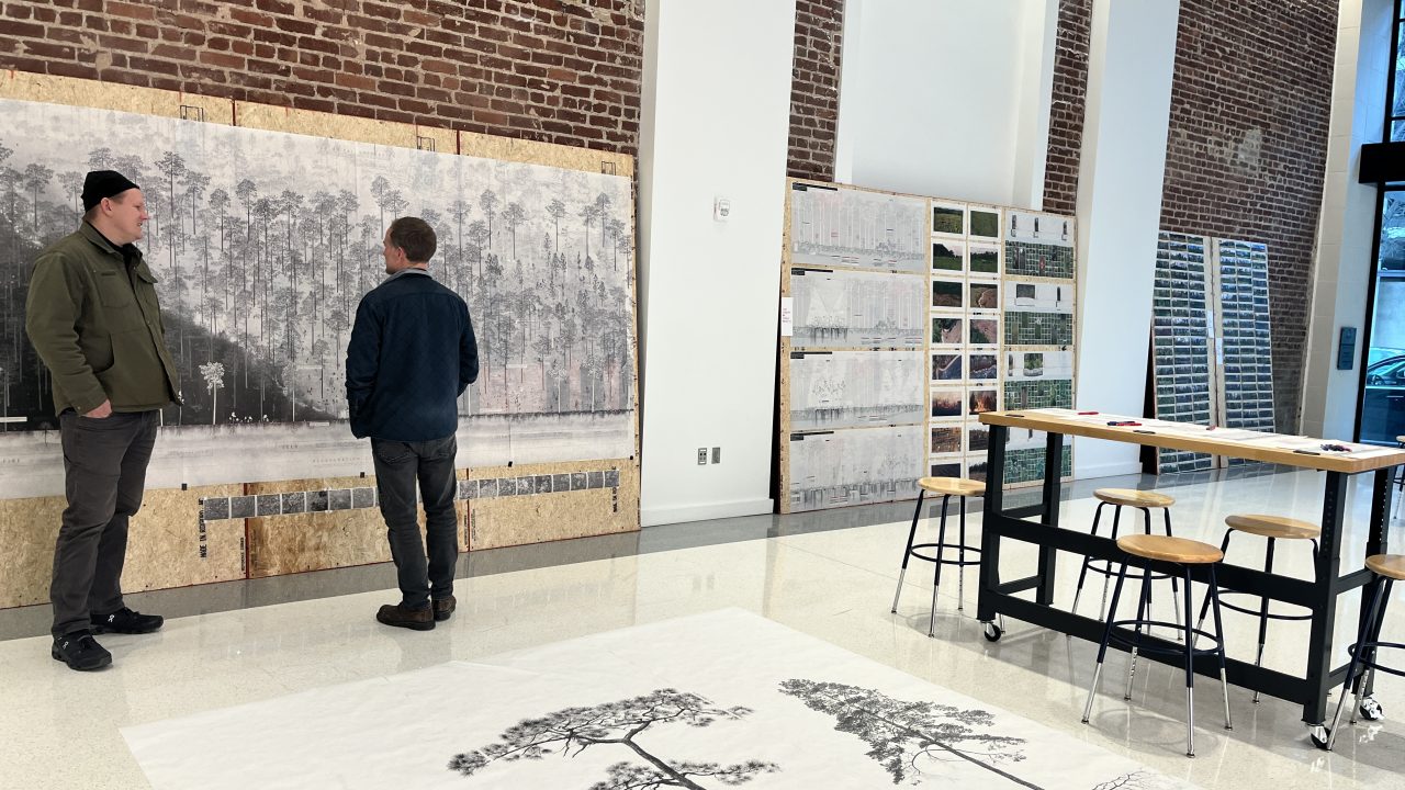 Two people stand in a modern studio space, looking at large black and white landscape prints on the wall, with tables, stools, and architectural drawings visible.