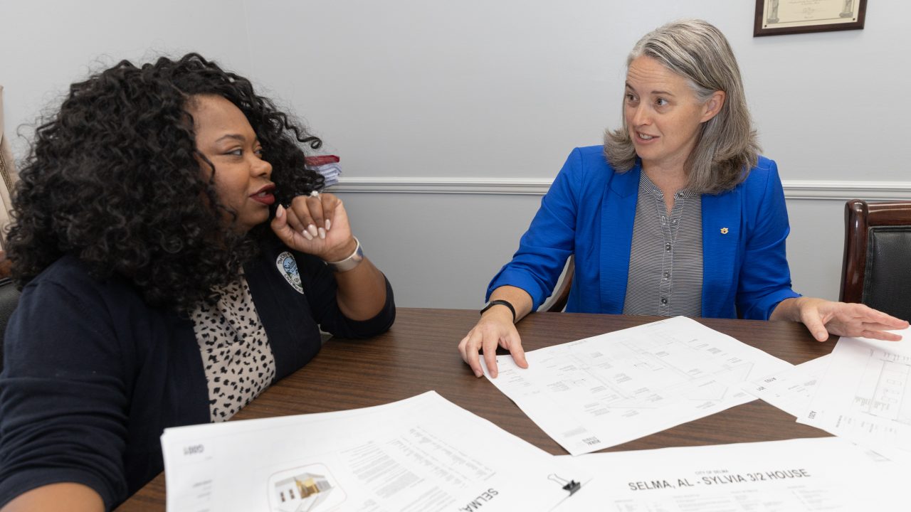 Two women sit at a table discussing architectural plans and documents in an office setting.