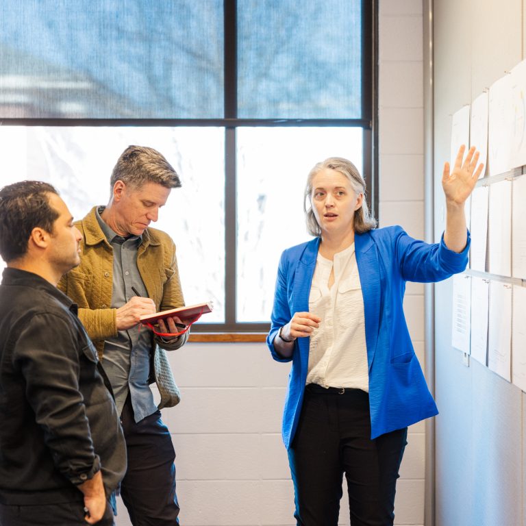 A woman in a blue blazer presents charts on a wall to two colleagues, one of whom is taking notes on a tablet.