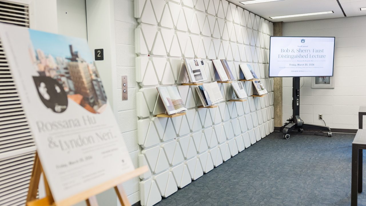 A hallway with a display of books and magazines on shelves, a digital sign announcing the Rob & Sherry East Distinguished Lecture, and a poster for a Rossana Hu & Lyndon Neri event.