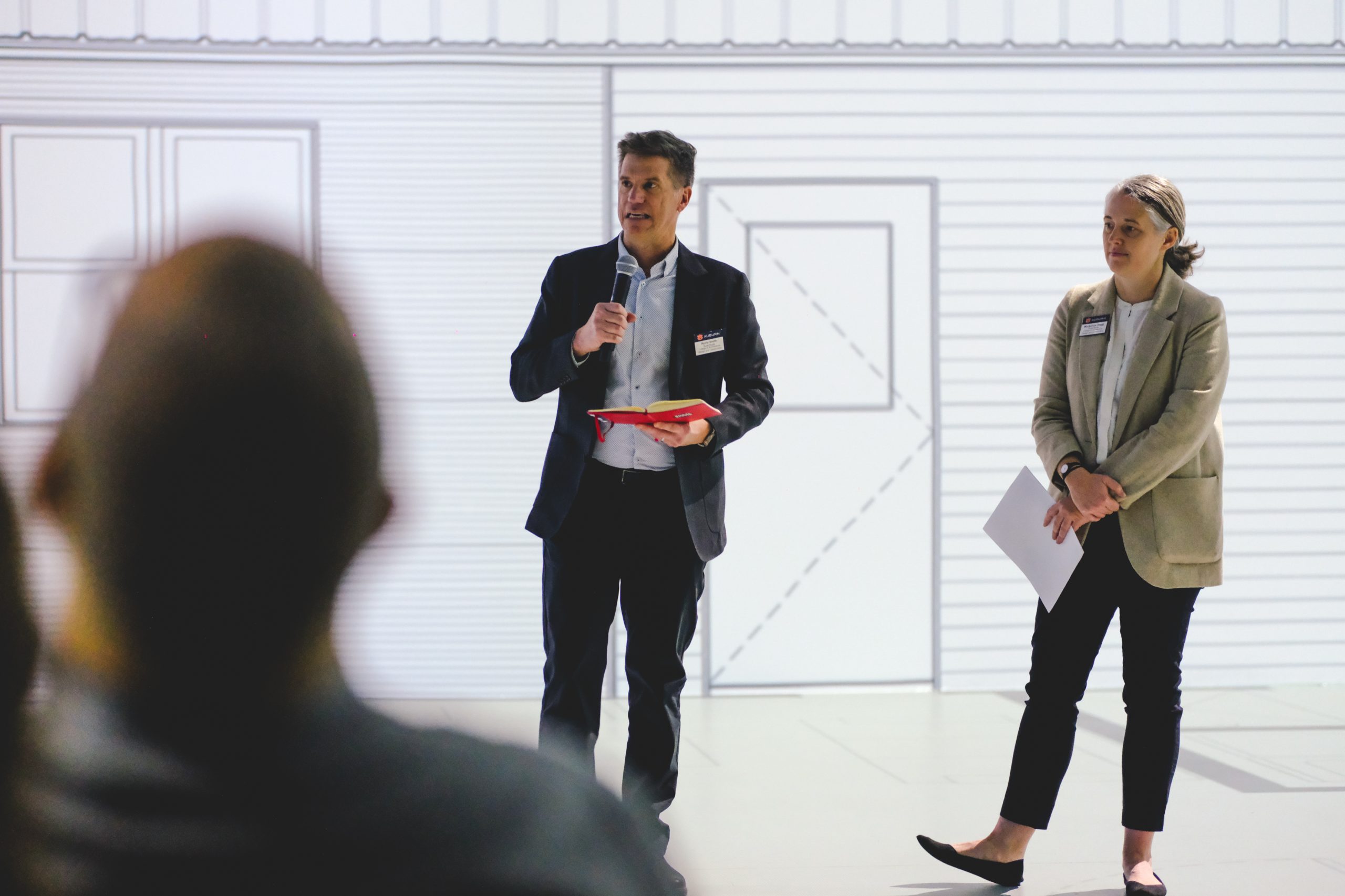 A man holding a microphone speaks to an audience while a woman stands beside him holding papers; both are wearing business attire.