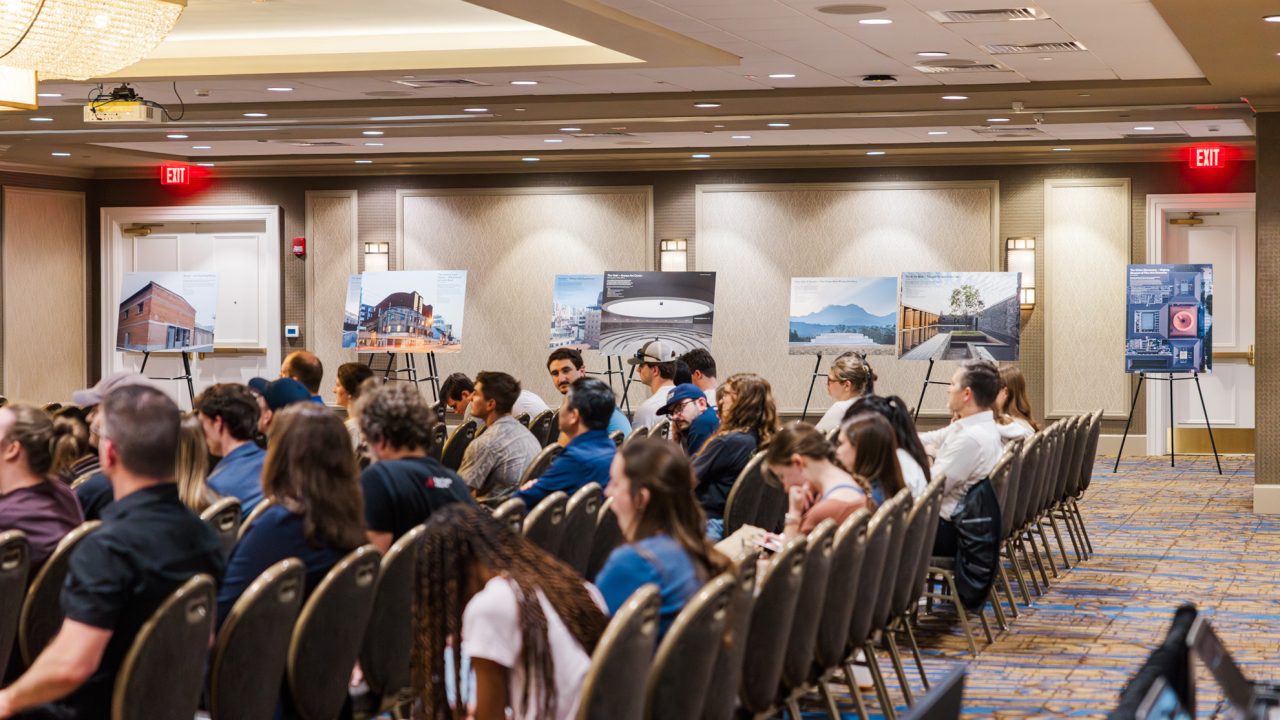 Audience seated in a conference room facing posters on easels displaying architectural designs and photographs along the back wall.