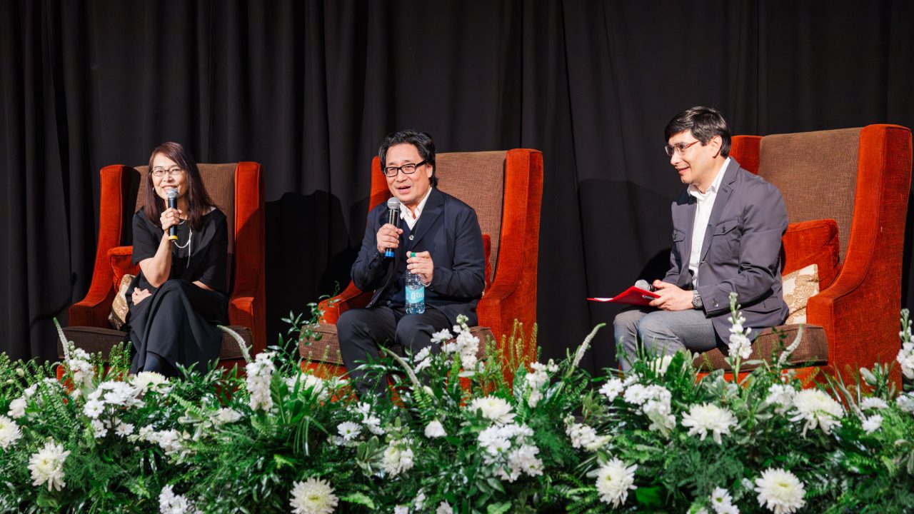 Three people sit on stage in high-backed chairs, speaking into microphones, with white floral arrangements in front and a black curtain backdrop.