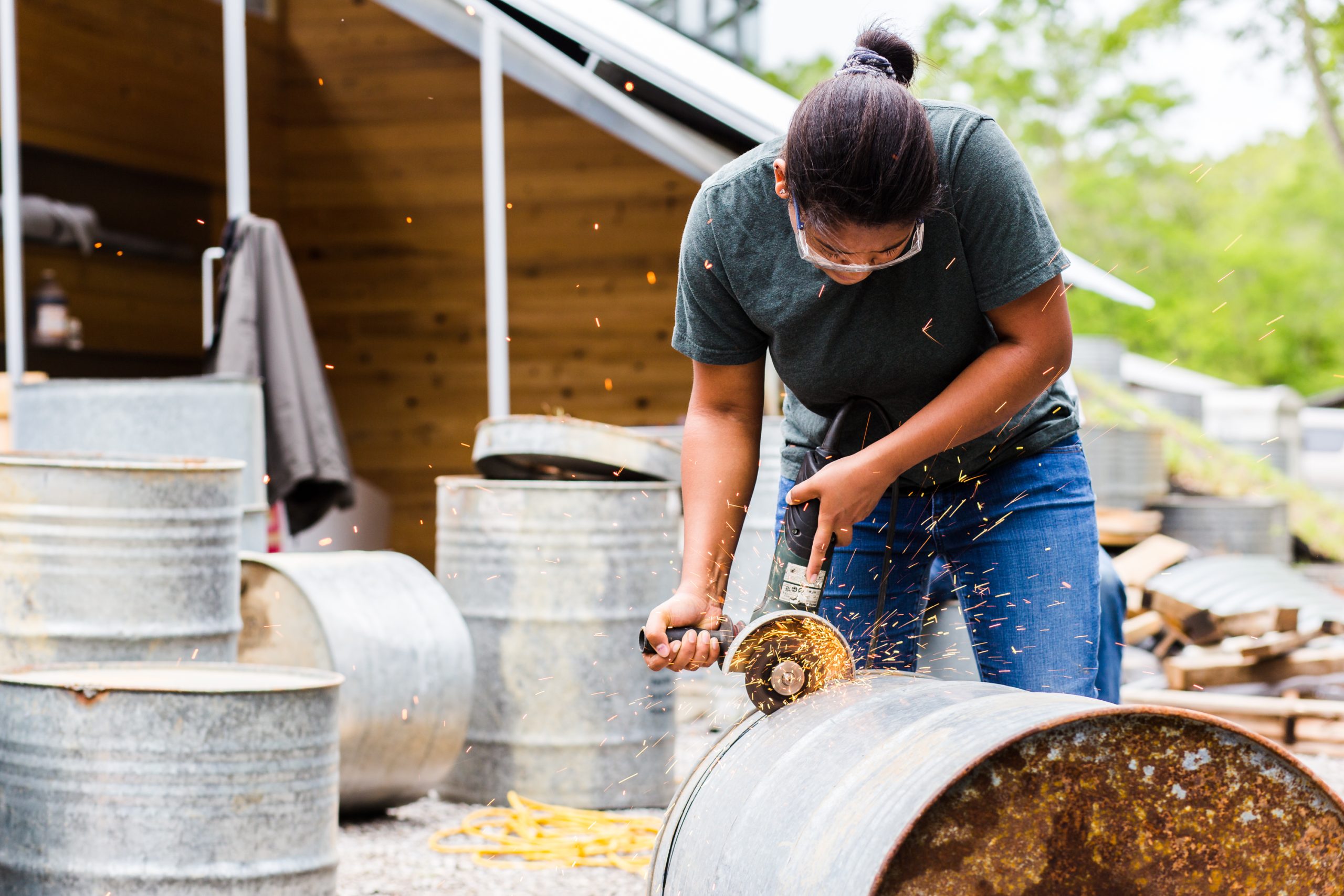 AEon McNeal uses a power grinder to cut a rusty metal barrel outdoors, with sparks flying and protective eyewear on. Other barrels and a wooden structure are in the background.