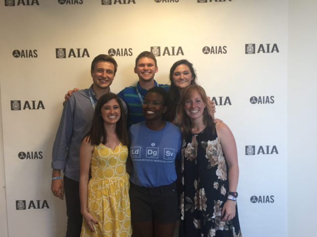 Six people stand smiling in front of a backdrop printed with the AIA and AIAS logos.