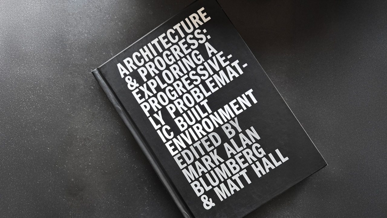 A black hardcover book titled Architecture & Progress: Exploring a Progressively Problematic Built Environment, edited by Mark Alan Blumberg and Matt Hall, on a dark surface.