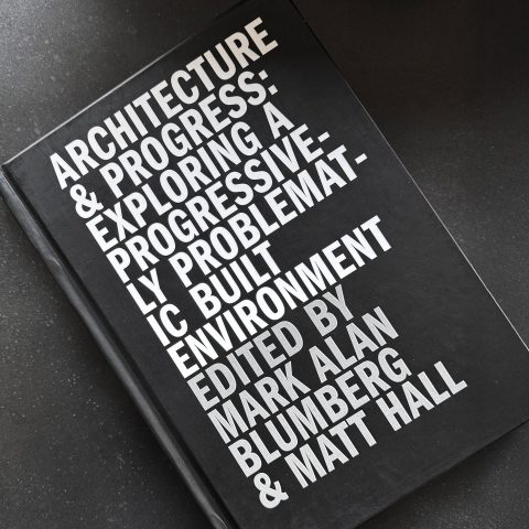 A black hardcover book titled Architecture & Progress: Exploring a Progressively Problematic Built Environment, edited by Mark Alan Blumberg and Matt Hall, on a dark surface.