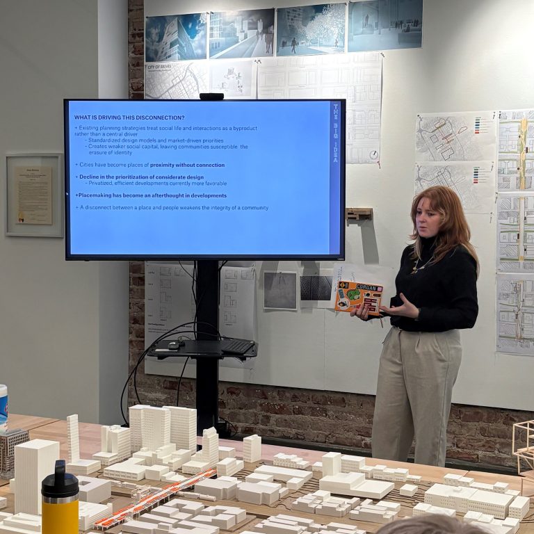 A woman presents to a group in a design studio, standing beside a screen displaying presentation slides and architectural models on the table.