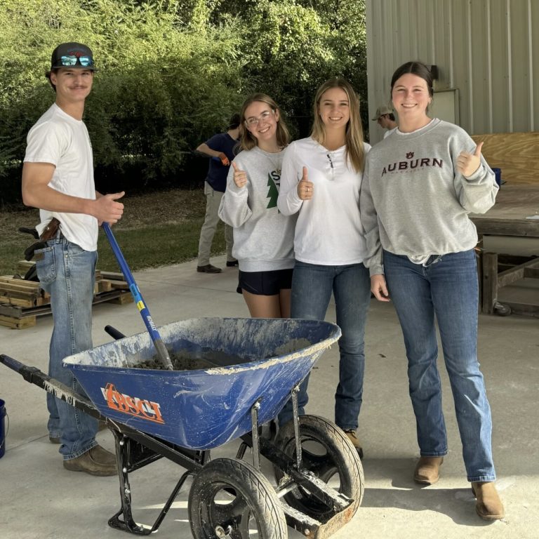 Four young adults stand by a wheelbarrow with cement, smiling and giving thumbs up, outside a building near some construction materials.