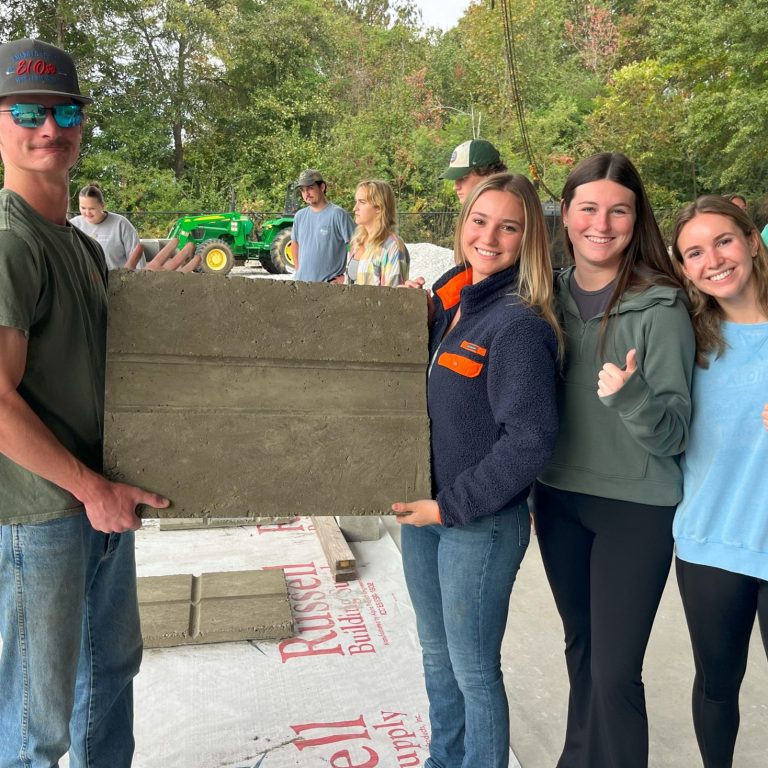 Four young adults stand outdoors; two hold a large rectangular slab of wet concrete while the other two smile and give a thumbs up. Other people and a green tractor are visible in the background.