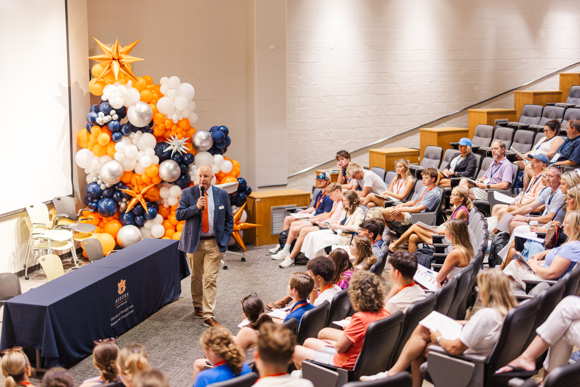 A man with a microphone speaks to an audience seated in a lecture hall; a balloon arrangement decorates the stage area.