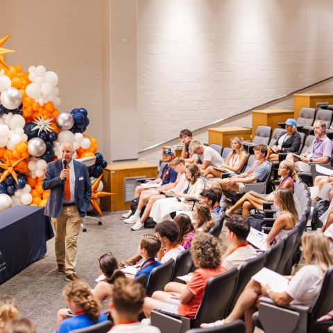 A man with a microphone speaks to an audience seated in a lecture hall; a balloon arrangement decorates the stage area.