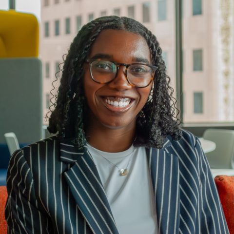 A person with glasses and curly hair, wearing a striped blazer, sits on an orange couch in a modern office space with large windows and yellow partitions.