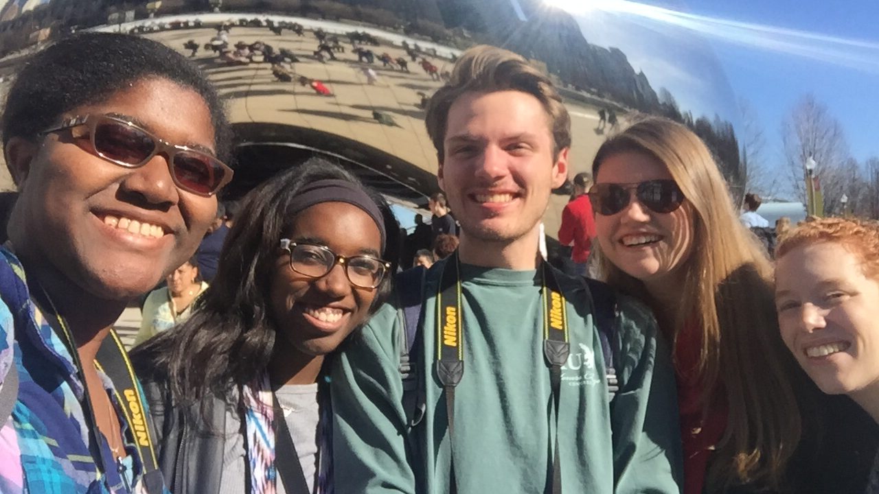 Five people smiling for a selfie in front of the reflective Cloud Gate sculpture (The Bean) in Millennium Park, Chicago, on a sunny day.