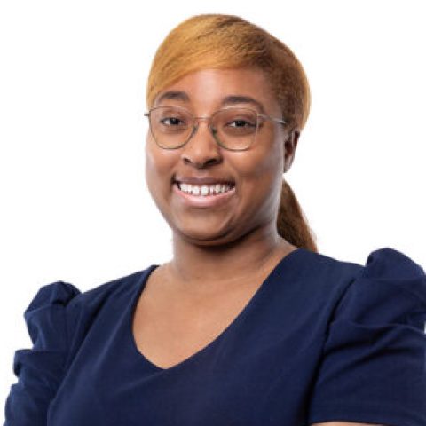 A woman with glasses and light brown hair in a ponytail, wearing a navy blue dress, stands smiling with her hands folded in front of her against a white background.