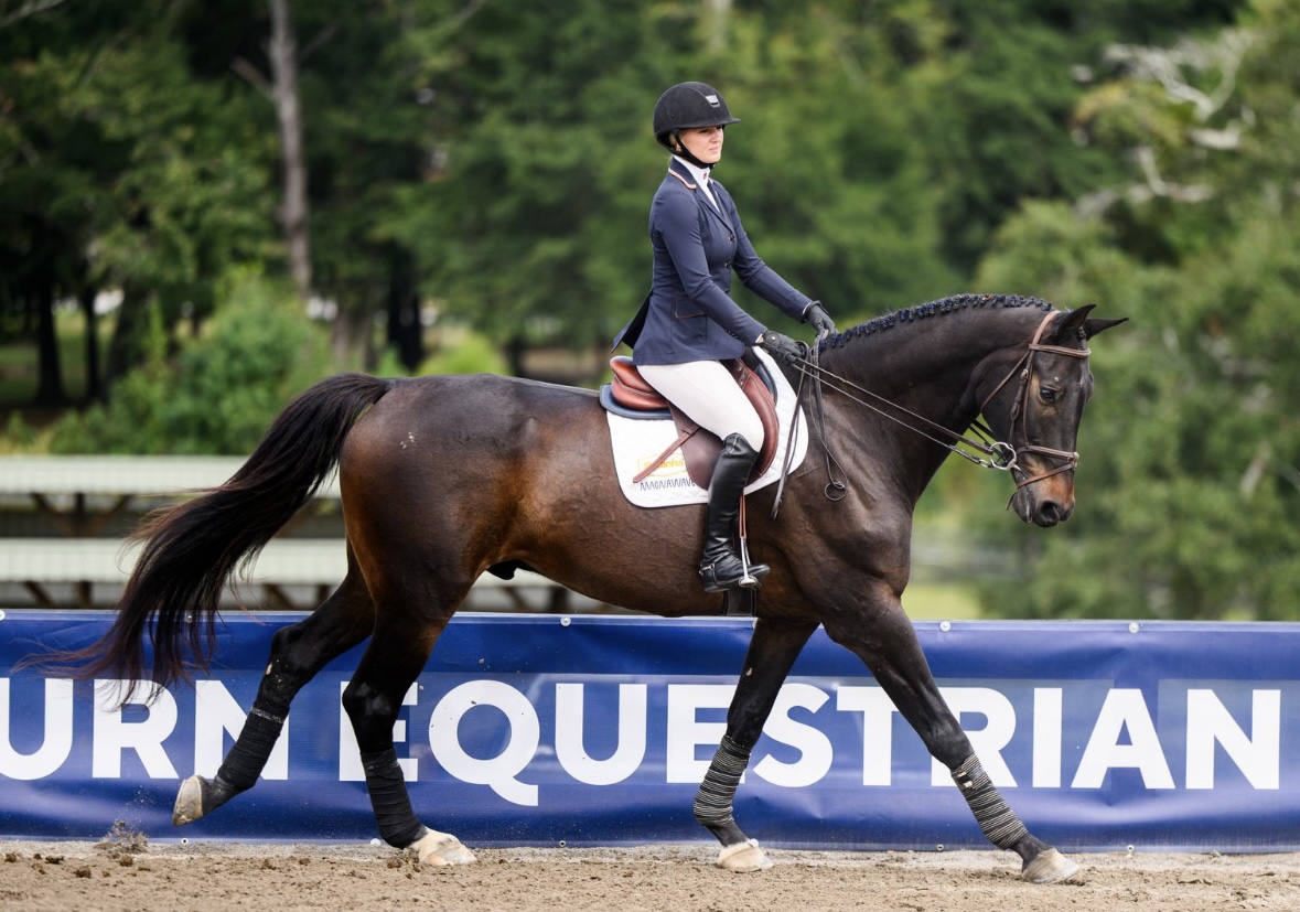 A person in equestrian attire rides a dark brown horse during a dressage event, with a blue EQUESTRIAN sign in the background and trees behind.