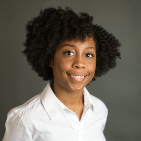 A woman with short curly hair smiles while wearing a white button-down shirt and black skirt, standing in front of a plain gray background.