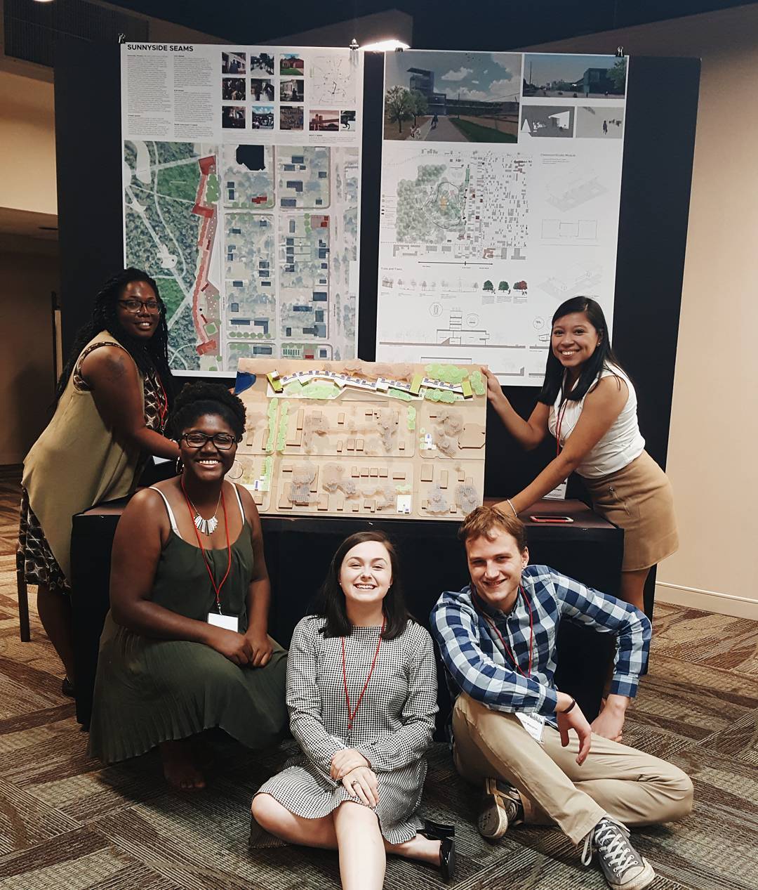 Five people pose in front of an architectural project display featuring maps, diagrams, and a scale model on a table.