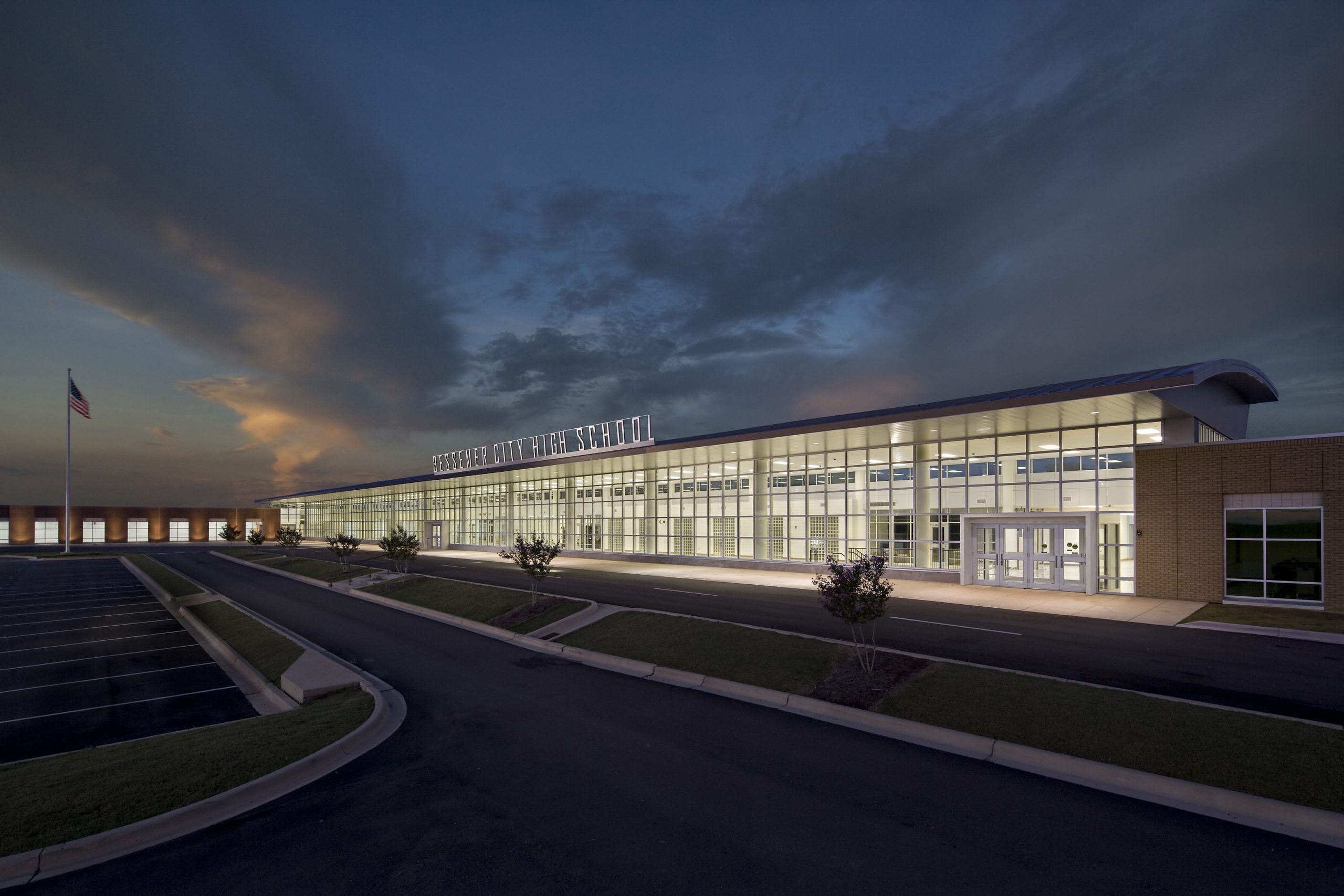 A modern high school building with large glass windows is lit up at dusk, with an empty parking lot and an American flag on the left.