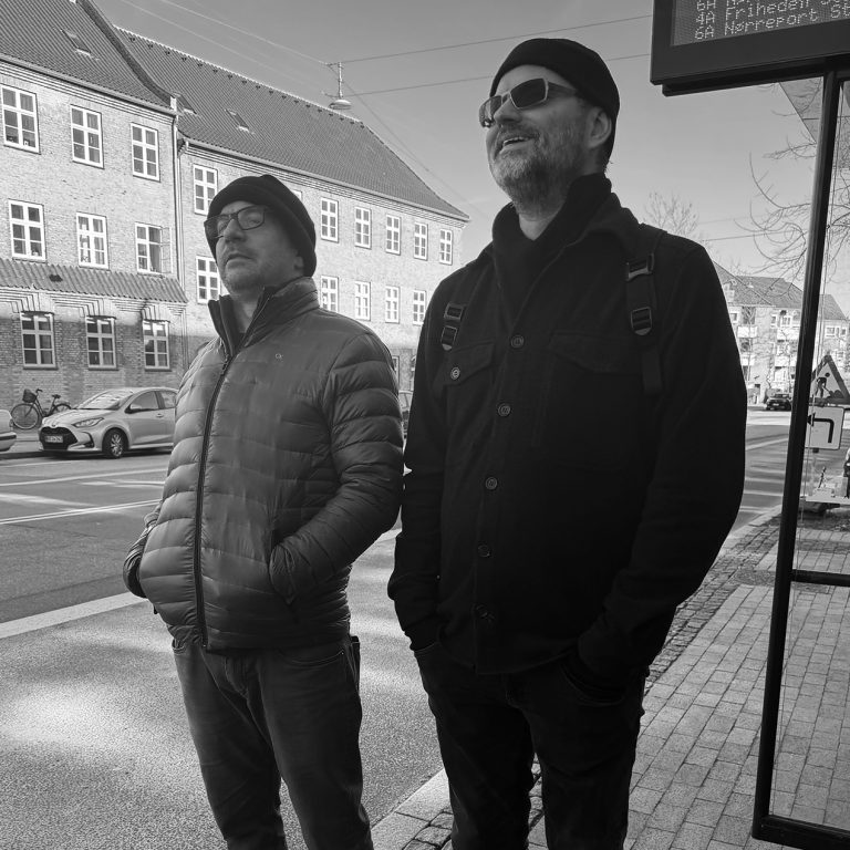 Two men wearing jackets and hats stand on a sidewalk near a bus stop, with buildings and a street in the background. The photo is in black and white.