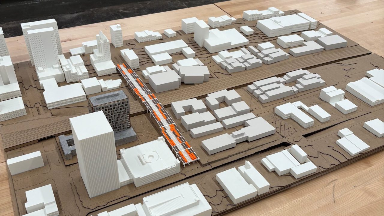 Architectural scale model of an urban area with white building blocks, orange road markings, and city layout on a wooden table. Blueprints are visible on the wall in the background.