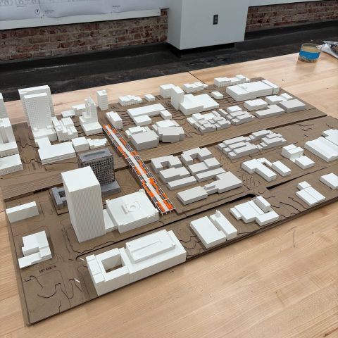 Architectural scale model of an urban area with white building blocks, orange road markings, and city layout on a wooden table. Blueprints are visible on the wall in the background.