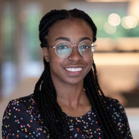 A woman with glasses and long braids smiles, wearing a black, patterned blouse in a blurred indoor office setting.