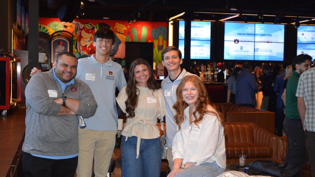 Five people pose and smile for a group photo at an indoor event, with a bar and crowd in the background. Four are standing and one is seated in front.