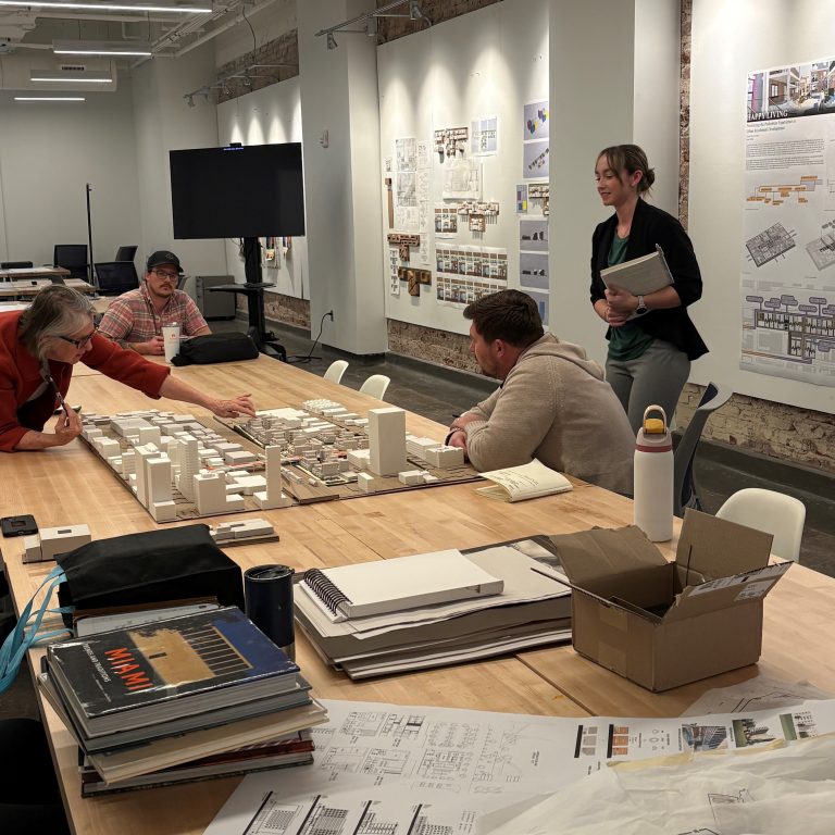 A group of people reviews architectural models and plans on a table in a modern office, while one person stands and presents near pinned-up design boards.