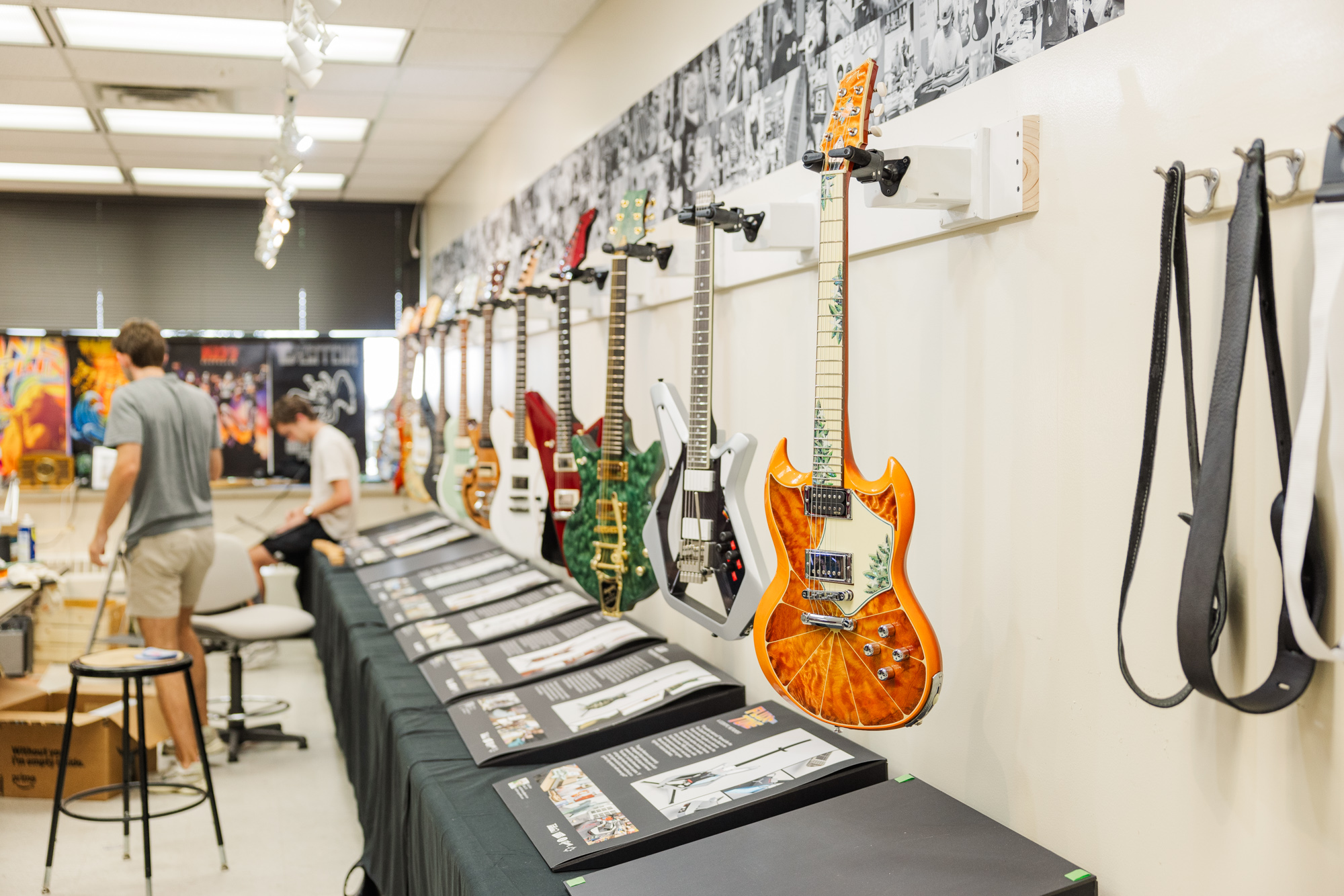 A row of electric guitars hangs on a wall above display tables with printed materials; two people work in the background of a well-lit room.