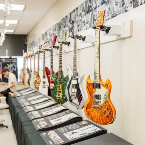 A row of electric guitars hangs on a wall above display tables with printed materials; two people work in the background of a well-lit room.