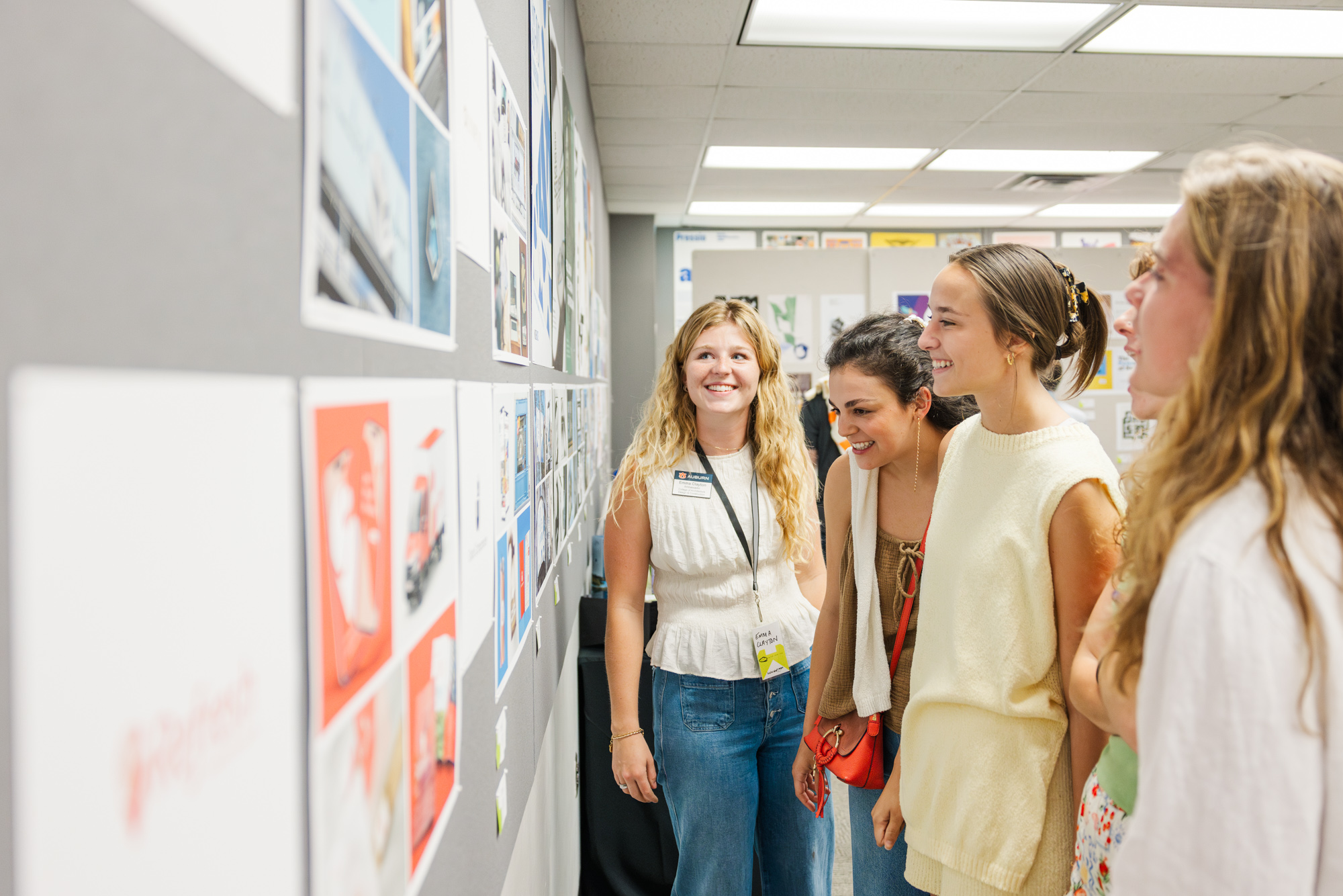 Four young women stand together, looking at artwork or project displays pinned to a gray bulletin board in a well-lit room.
