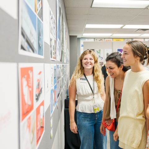 Four young women stand together, looking at artwork or project displays pinned to a gray bulletin board in a well-lit room.