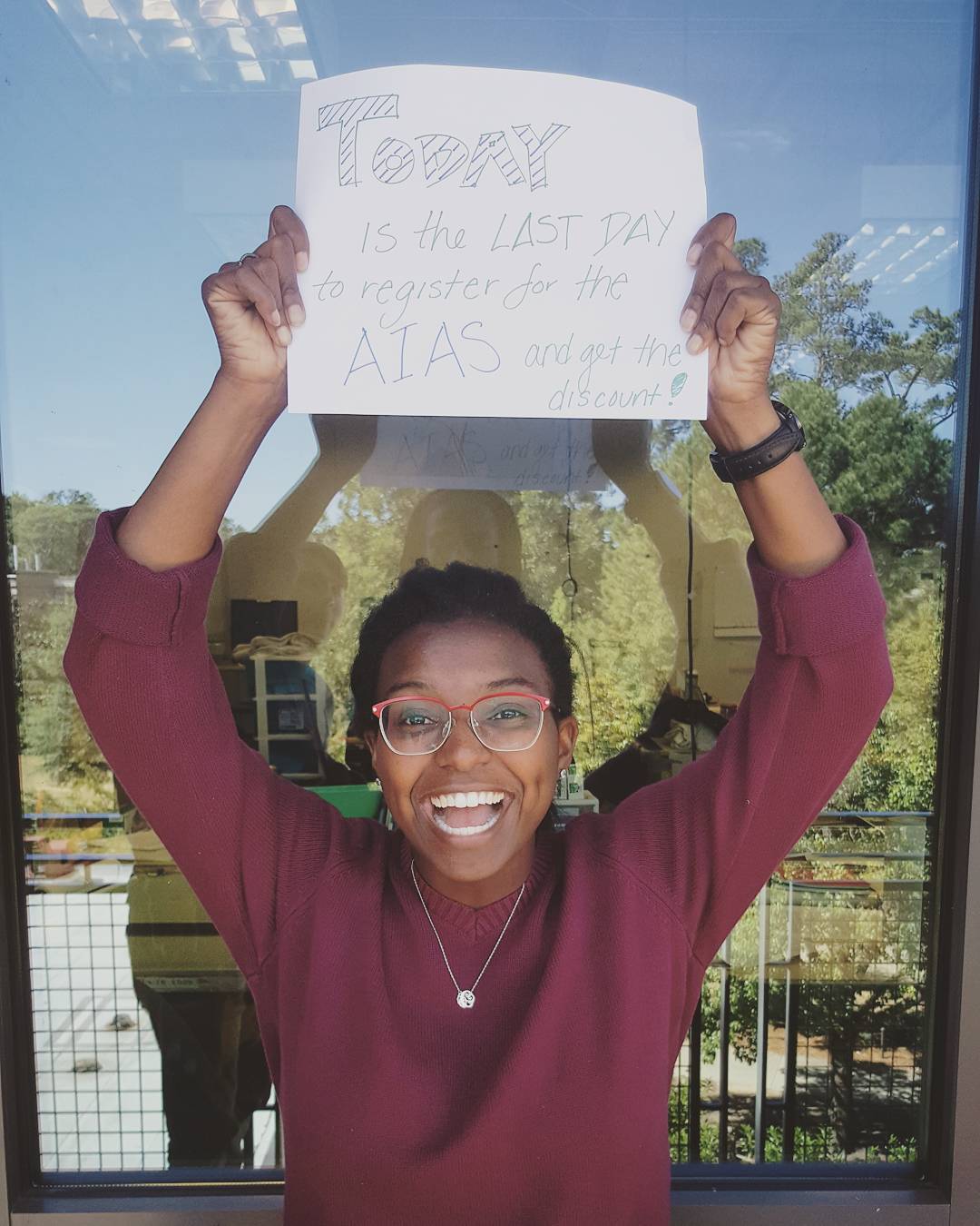 A smiling person holds up a sign that says, Today is the last day to register for the AIAS and get the discount, standing in front of a window with trees outside.