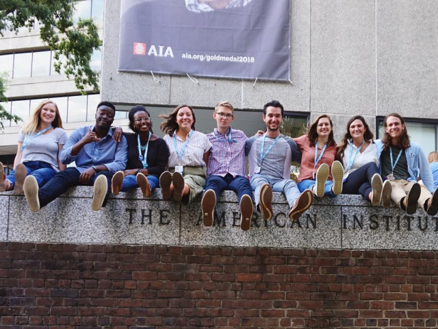 Nine people sit side by side on a stone ledge outside a building, smiling at the camera, with their legs extended forward and name badges around their necks.