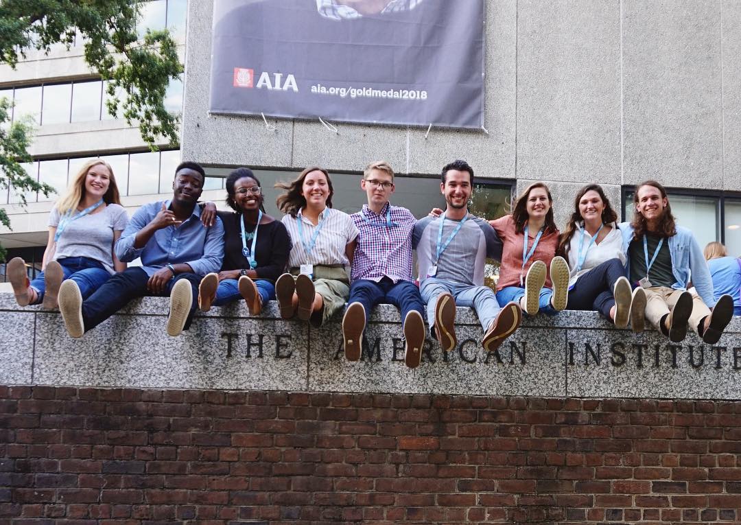 Nine people sit side by side on a stone ledge outside a building, smiling at the camera, with their legs extended forward and name badges around their necks.