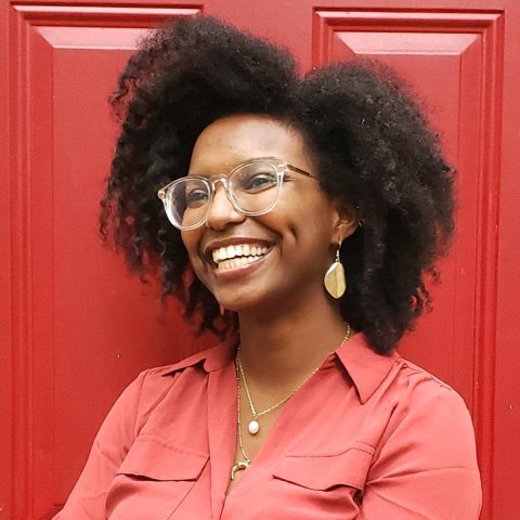 Person with curly hair, wearing glasses and a pink button-up shirt, stands smiling with arms crossed in front of a red door.