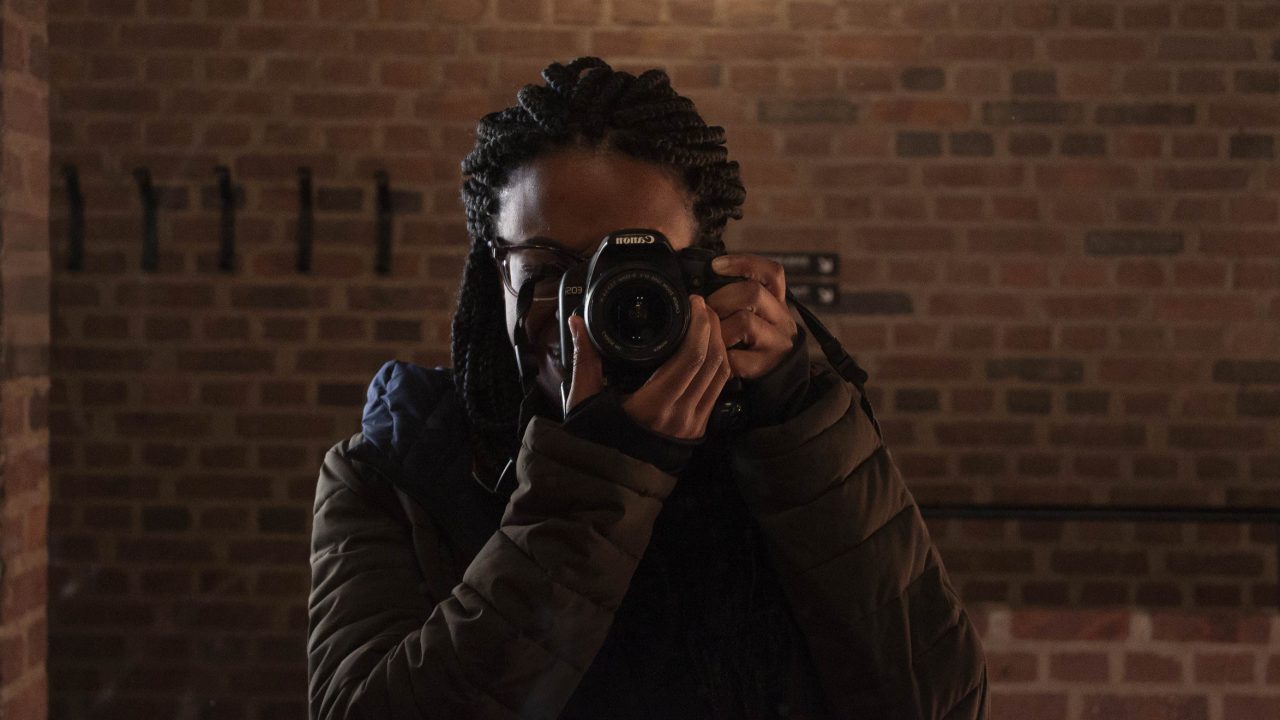 A person with braided hair and glasses takes a photo with a camera in front of a brick wall, under a bright light.